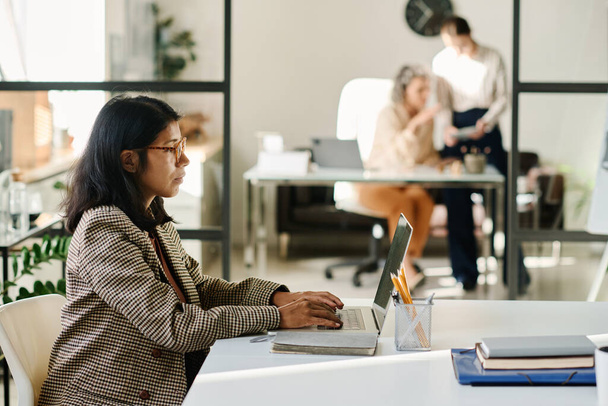 Laptop ekranındaki çevrimiçi verilere bakan genç kadın analistin yan görünümü ve birlikte çalışma alanında otururken klavyede daktilo yazması - Fotoğraf, Görsel