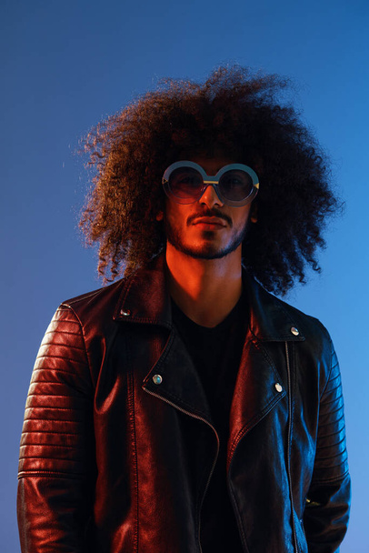 Portrait of a stylish man with curly hair with glasses smile on blue background multinational, color light, black leather jacket trend, modern concept. High quality photo - Φωτογραφία, εικόνα