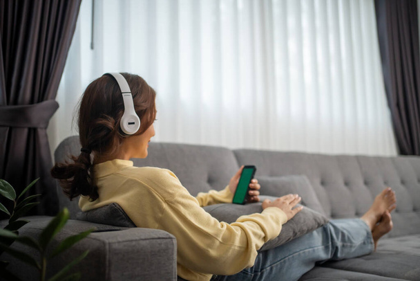 Asijka sedět na pohovce v obývacím pokoji doma, aby si jejich oblíbené písně. dívka nosí sluchátka poslouchat hudbu, náladu a hobby, moderní bezdrátové technologie koncept - Fotografie, Obrázek