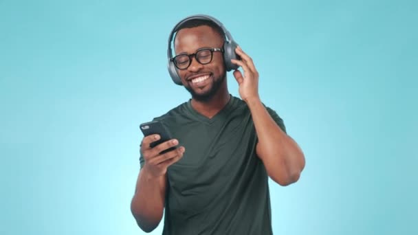 Fejhallgató, telefon és fiatal fekete férfi egy stúdióban zenét hallgatni, albumot vagy lejátszási listát. Happy, tánc és afrikai férfi modell streaming egy dalt vagy rádiót mobiltelefon elszigetelt kék háttér - Felvétel, videó