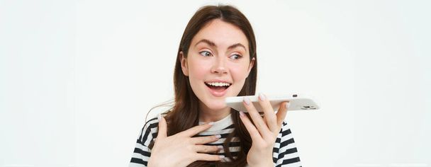 Image of cute brunette woman talking into speakerphone, holding mobile phone near mouth, records her voice, sends a voicemessage, using online translator app, white background. - Photo, Image
