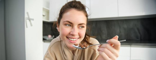 Image d'une jeune femme heureuse mangeant des céréales à la maison avec du lait, prenant son petit déjeuner, portant un peignoir, assise dans la cuisine seule. - Photo, image