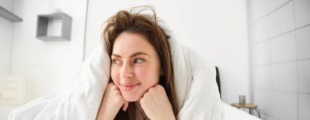 Roztomilá dívka s rozcuchanými vlasy, ležící v posteli přikrytá bílou přikrývkou, usmívající se a smějící se koketka, trávící čas ve své ložnici. - Fotografie, Obrázek