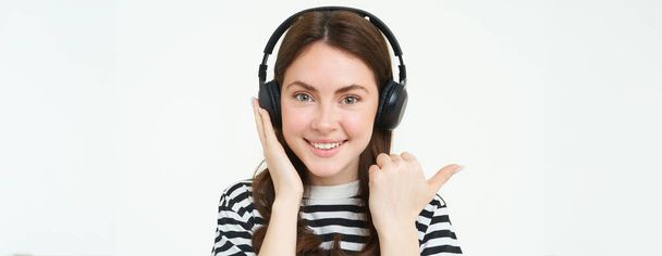 Musikspeicherkonzept. Lächelnde junge Frau hört Song über Kopfhörer, zeigt links auf Kopierraum, zeigt Werbetext, steht über weißem Hintergrund. - Foto, Bild