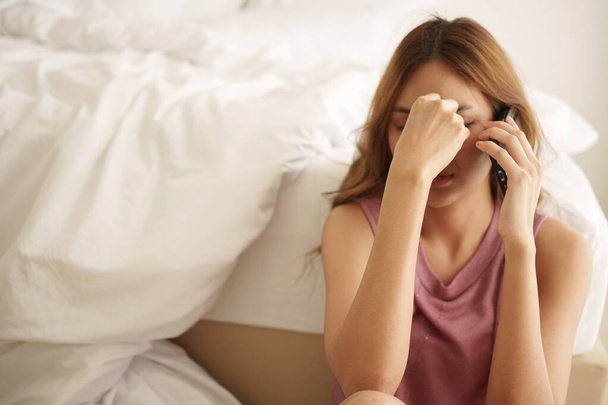 Нещаслива азіатська жінка сидить на підлозі спальні, покриваючи обличчя рукою і використовуючи смартфон з тривожним виразом в спальні. - Фото, зображення