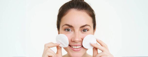 Portret młodej kobiety myjącej twarz, zmywającej makijaż podkładkami z bawełny, używając tonera do twarzy, środka do czyszczenia skóry, stojącej nad białym tłem. - Zdjęcie, obraz