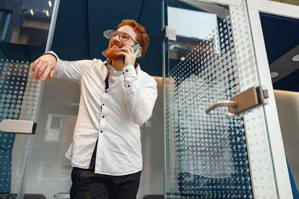 Uśmiechnięty menadżer w okularach rozmawia przez telefon stojący w nowoczesnej przestrzeni dla współpracowników - Zdjęcie, obraz
