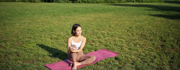 Kobieta w parku, ogląda film z jogi na smartfonie, medytuje na świeżym powietrzu, siedzi na gumowej macie. - Zdjęcie, obraz