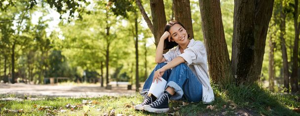 Nainen istuu puistossa suosikkikirjansa kanssa, nojaten puuhun aurinkoisena päivänä, nauttien luonnosta ja rauhallisesta rentouttavasta ilmapiiristä.. - Valokuva, kuva