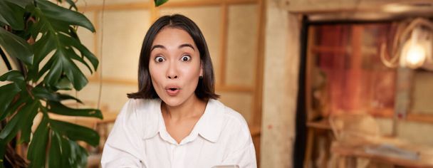 Retrato de menina asiática olhando surpreso, segurando telefone celular com rosto surpreso, grande notícia no smartphone, sentado no café com livro. - Foto, Imagem