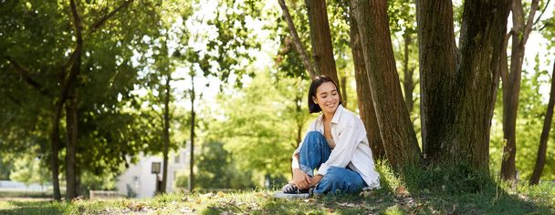 Muotokuva kaunis aasialainen nainen lepää lähellä puuta, rentouttava puistossa, hymyilee ja näyttää onnelliselta. Kopioi tila - Valokuva, kuva