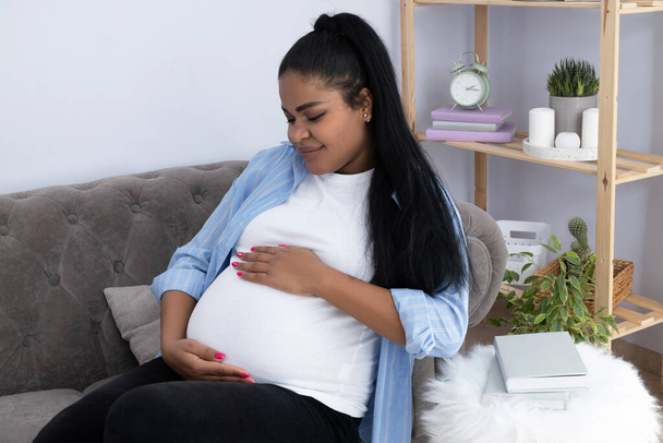 Mujer afroamericana embarazada en la habitación en el sofá. - Foto, imagen