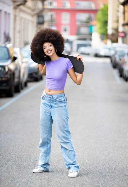 Corps complet de jeune femme afro-américaine positive avec des cheveux bouclés posant avec skateboard sur la rue de la ville - Photo, image