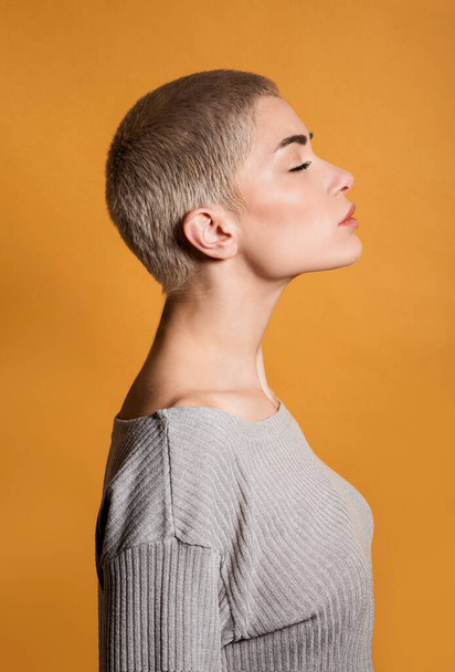 Profile of side view of thoughtful young female blonde with short hair and closed eyes standing in room with chin up against orange wall - Фото, изображение