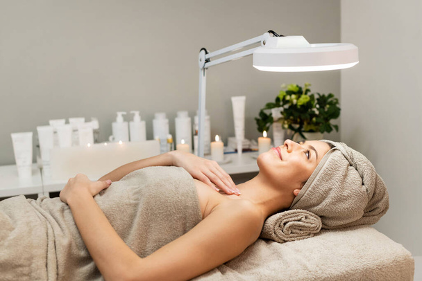 Widok z boku zachwycona kobieta z ręcznikami na głowę i ciało leżące na łóżku pod lampą w centrum urody - Zdjęcie, obraz
