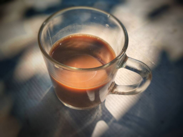 Επιλεκτική εστίαση του καφέ γάλα με θολή φόντο. ένα διάφανο φλιτζάνι καφέ γάλακτος στις πρωινές σκιές και ακτίνες του ήλιου μέσα από το παράθυρο. Πρωινή ιδέα καφέ, κοντινό πλάνο - Φωτογραφία, εικόνα