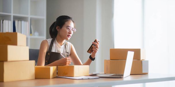 Serbest çalışan Asyalı kadın girişimcisi akıllı telefon ve kutu kullanarak müşterilere ve çevrimiçi iş fikirlerine satış hazırlamak için çevrimiçi sipariş alıyor ve gözden geçiriyor.. - Fotoğraf, Görsel