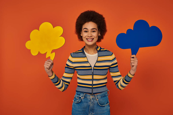 jovem mulher afro-americana posando com bolhas de pensamento azul e amarelo no fundo laranja - Foto, Imagem