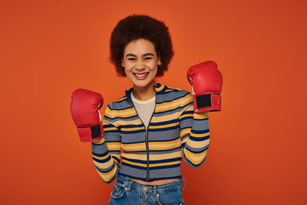 beautiful jolly african american woman posing actively with boxing gloves on orange backdrop - Zdjęcie, obraz