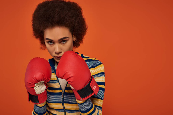 beautiful focused african american woman posing actively with boxing gloves on orange backdrop - Photo, Image