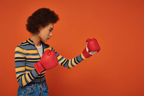 オレンジ色の背景にボクシング手袋で積極的にポーズする美しいスポーティなアフリカ系アメリカ人女性 - 写真・画像