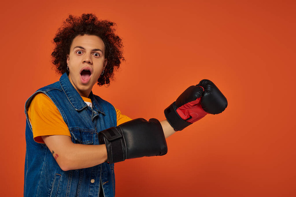 modelo masculino afroamericano deportivo sorprendido posando animado en guantes de boxeo sobre fondo naranja - Foto, imagen