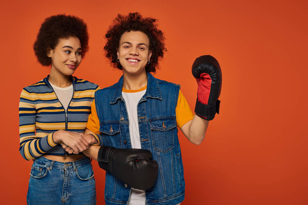 joyous african american man in boxing gloves having fun with his sister on orange backdrop, family - Photo, Image