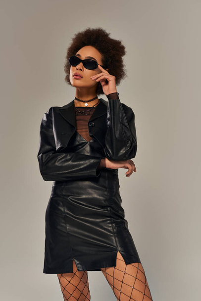 séduisante femme afro-américaine élégante en tenue noire avec des lunettes de soleil posant sur fond gris - Photo, image