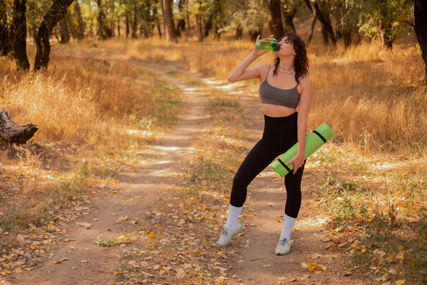 Жінка п'є воду з пляшки, стоячи в осінньому парку, після фітнес-тренувань, тримає йога килимок, концепція здорового зволоження в тренувань - Фото, зображення