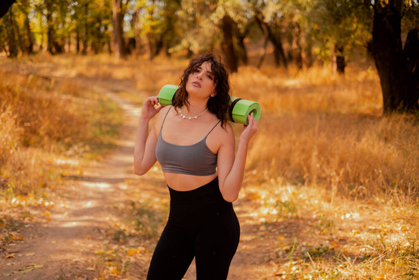 Kobieta pijąca wodę z butelki stojąc w jesiennym parku, po treningu fitness, trzymając matę do jogi, koncepcja zdrowego nawilżenia podczas treningów - Zdjęcie, obraz