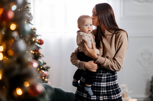 Szczęśliwa młoda matka trzyma swojego synka w ramionach na tle pięknie zdobionego Nowego Roku drzewa i całuje go. Świąteczna rodzinna atmosfera. Piękny portret matki i syna - Zdjęcie, obraz