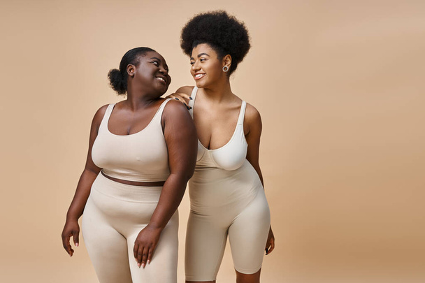 felice plus size donne africane americane in lingerie sorridente a vicenda sul beige, positività del corpo - Foto, immagini