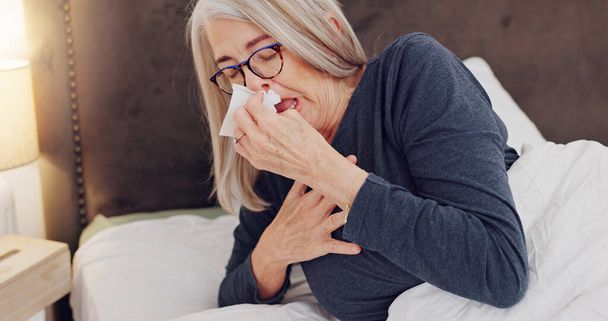 Άρρωστος, φυσάει μύτη και ηλικιωμένη γυναίκα στο κρεβάτι με αλλεργίες, γρίπη ή κρύο το πρωί του Σαββατοκύριακου στο σπίτι. Ασθένεια, φτάρνισμα και ηλικιωμένη γυναίκα σε συνταξιοδότηση με ιστό για κόλπων στο υπνοδωμάτιο στο σπίτι - Φωτογραφία, εικόνα