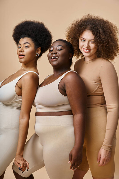 charming multiethnic plus size women in lingerie smiling at camera on beige backdrop, body positive - Photo, Image