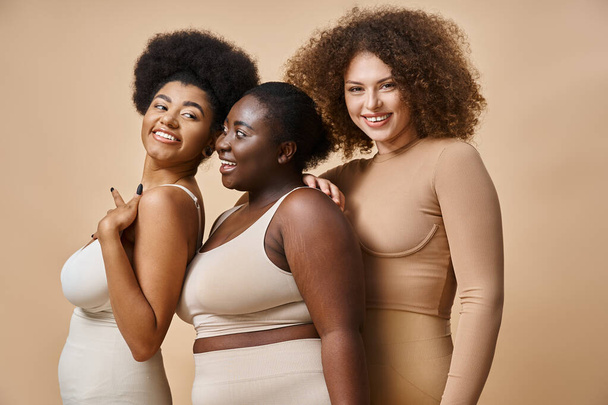 fröhliche multiethnische Plus-Size-Frauen in Unterwäsche posieren vor beigem Hintergrund, Selbstbewusstsein und Charme - Foto, Bild