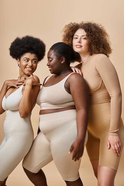 joyful multiethnic plus size women in underwear posing on beige backdrop, self-confidence and charm - Zdjęcie, obraz