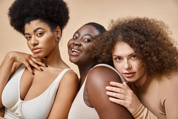 fröhliche Plus-Size-Frau in Dessous blickt in die Kamera neben multiethnischen Freundinnen auf beige - Foto, Bild