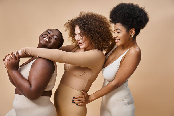 allegro multiculturale plus size donne in lingerie abbracciando su beige, positività del corpo e bellezza - Foto, immagini