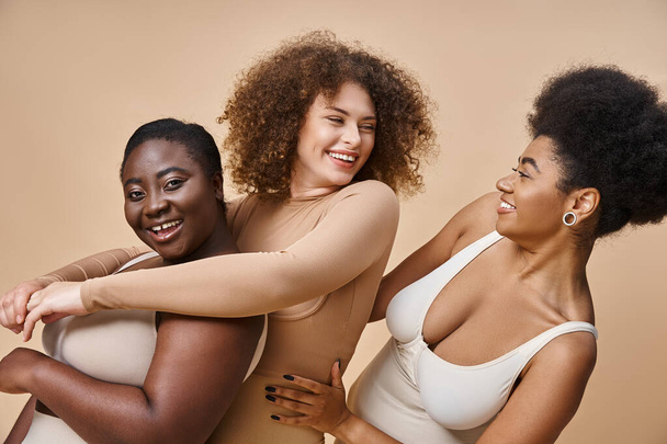 χαρούμενα πολυπολιτισμική συν μέγεθος των γυναικών σε εσώρουχα αγκαλιάζει σε μπεζ, θετικότητα του σώματος και την ομορφιά - Φωτογραφία, εικόνα