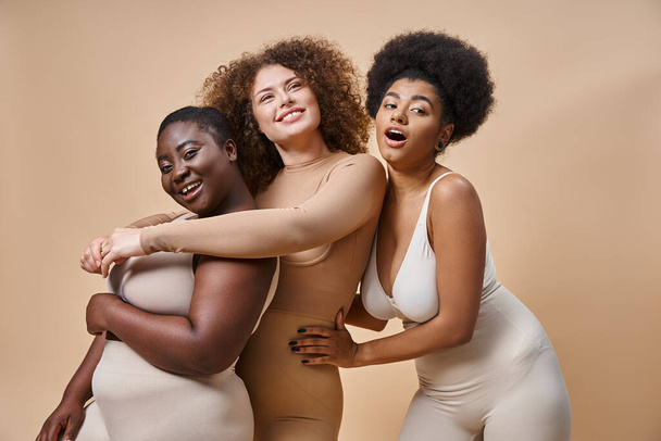 χαρούμενα πολυπολιτισμική συν μέγεθος των γυναικών σε εσώρουχα αγκαλιάζει σε μπεζ, θετικότητα του σώματος και την ομορφιά - Φωτογραφία, εικόνα