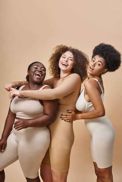 vzrušený multiraciální plus velikost ženy ve spodním prádle objímající a smějící se na béžové, tělo pozitivní - Fotografie, Obrázek