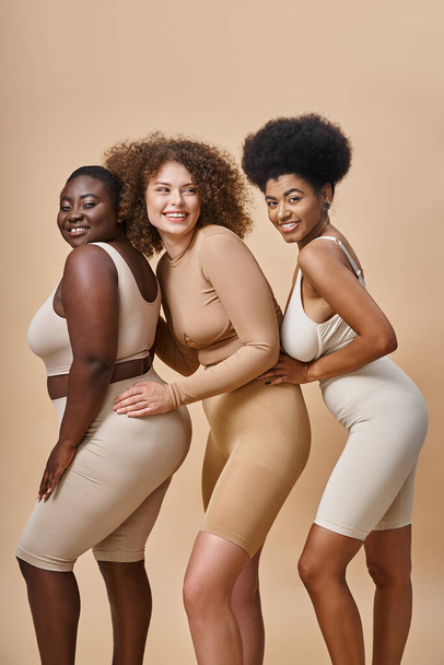 sonriente cuerpo positivo mujeres multirraciales en ropa interior posando sobre fondo beige, además de encanto tamaño - Foto, imagen