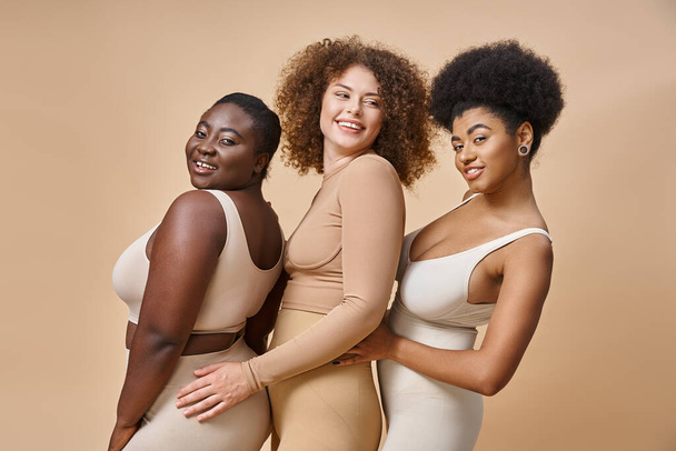 smiling body positive multiracial women in underwear posing on beige backdrop, plus size charm - Photo, Image