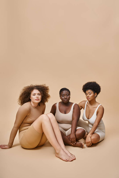 curvy and body positive multiethnic women in lingerie sitting and looking at camera on beige - Fotó, kép