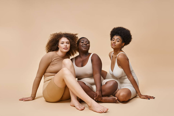 curvy and body positive multiethnic women in lingerie sitting and looking at camera on beige - Foto, immagini