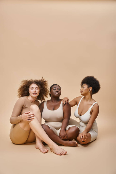 full length of multicultural plus size girlfriends in underwear sitting on beige, self-expression - Photo, Image
