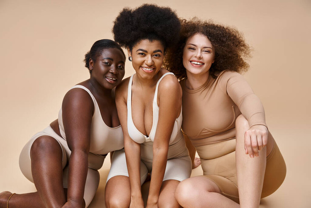 fröhliche multiethnische Plus-Size-Frauen in Unterwäsche lächeln auf beige, natürliche Körperschönheit - Foto, Bild