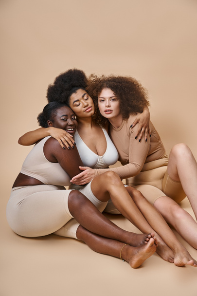 multiethnic plus size female friends in underwear sitting and embracing on grey, togetherness - Photo, Image