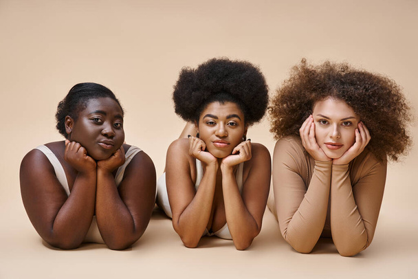 charmante corps multiethnique femmes positives en lingerie couché et regardant la caméra sur beige - Photo, image