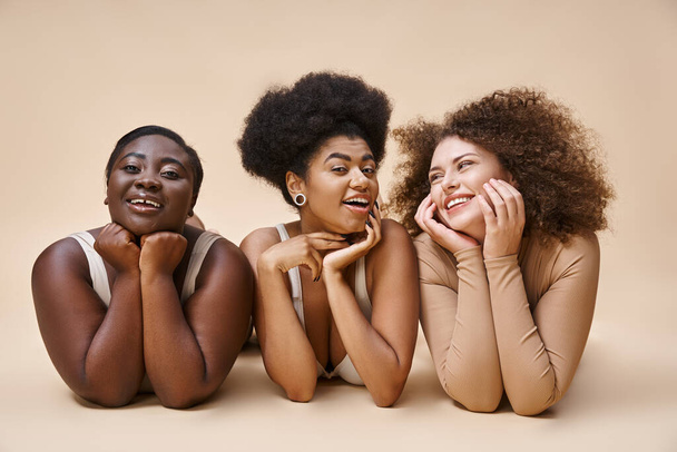 smiling multiethnic body positive women in lingerie lying down and posing on beige, natural beauty - 写真・画像